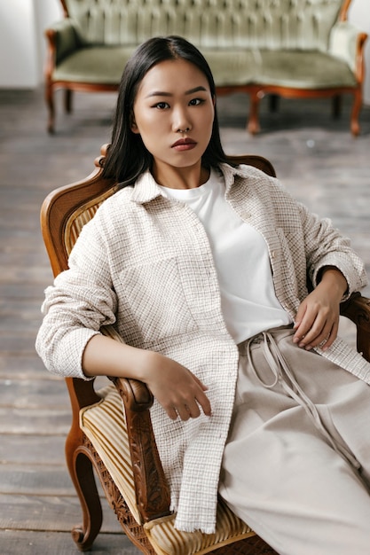 Browneyed brunette Asian woman in beige pants white tshirt and stylish cardigan sits on soft striped classic armchair