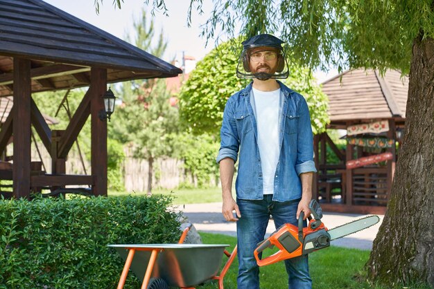 Brunet handyman in protective shield standing with cordless chain saw while working in backyard