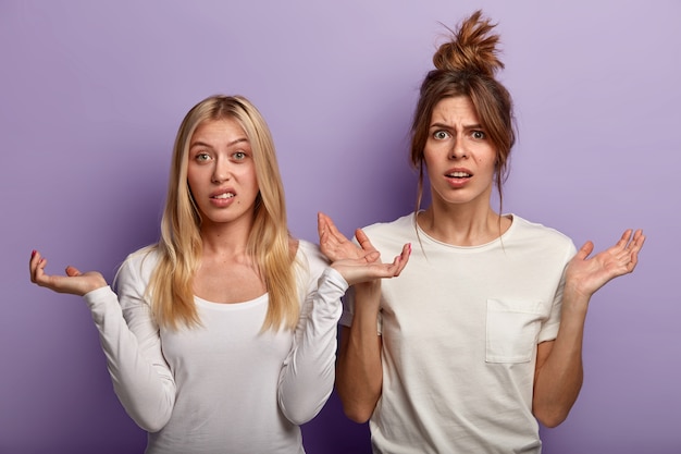 Free photo brunette and blonde women make confused uncertain gesture, shrug shoulders, should to decide something important, wear casual white clothes, isolated over purple wall. apathy, decision