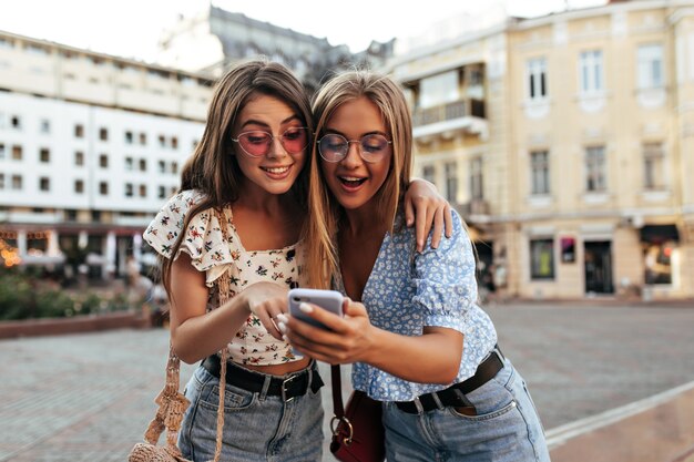 Brunette and blonde women in stylish outfits look surprised and read message in cellphone