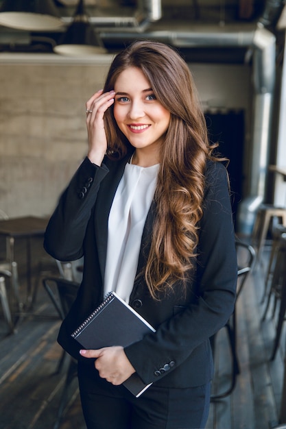 Free photo brunette businesswoman with wavy long hair and blue eyes stands holding a notebook in hands and smiles widely