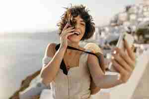 Free photo brunette curly woman in beige dress and straw hat smiles sincerely and takes selfie outside in old greek city happy lady in stylish outfit and boater holds phone on sea background