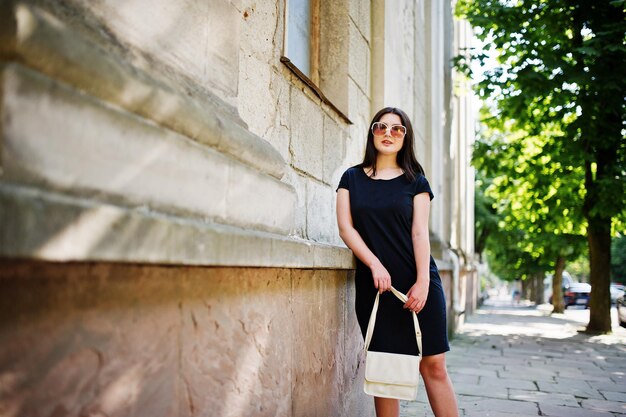 Brunette girl at black dress on sunglasses with handbag at hand posing at street of city