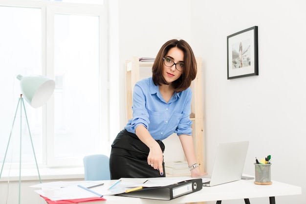 Brunette girl in blue shirt and black skirt is standing near table in office. She rested her hand on the table and shows to the papers . She is looking to the camera seriously.