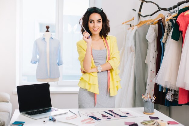 Free Photo a brunette girl in a gray dress and yellow jacket is standing near the table in a workshop studio. she has a lot of creative stuff  on the table . she is holding a pencil in a hand.