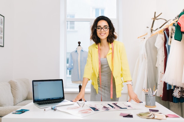 A brunette girl in a gray dress and yellow jacket is standing near the table in a workshop studio. She has a lot of creative stuff  on the table. She is smiling to the camera.