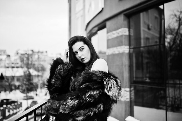 Brunette girl in green fur coat at street of city against house with large windows at winter