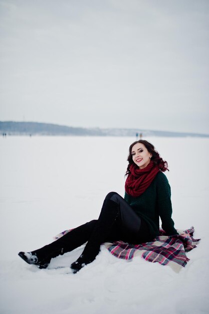 Brunette girl in green sweater and red scarf sitting on plaid outdoor frozen lake on evening winter day