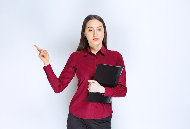 A brunette girl model standing with a folder and pointing away with an index finger.