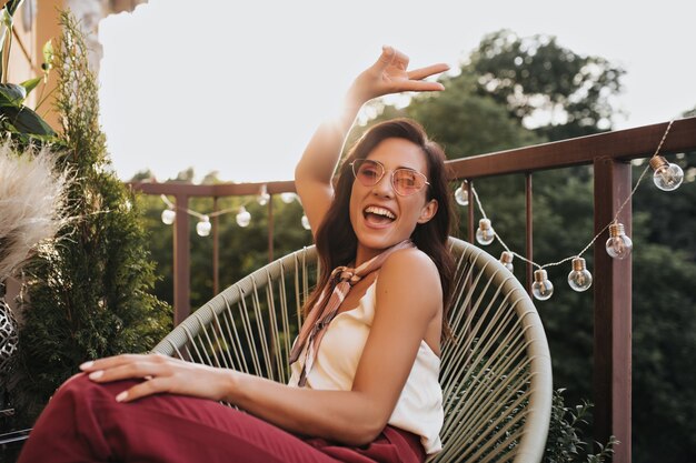 Brunette girl in pink glasses poses on balcony and shows peace sign. Cheerful woman in silk scarf around her neck laughs and sits on terrace.