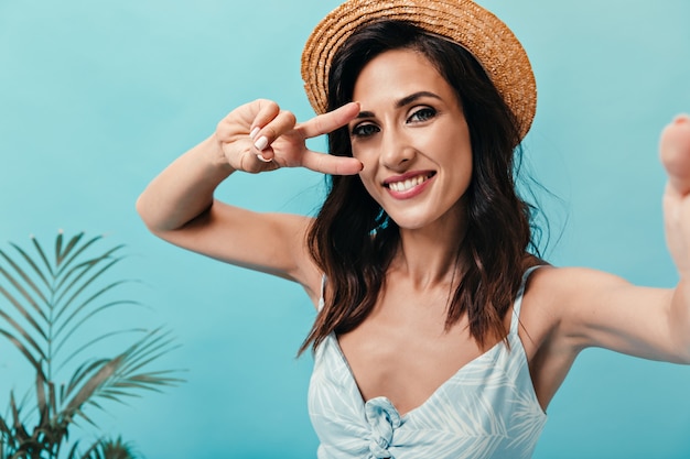 Brunette girl in straw hat shows peace sign and takes selfie on blue background. Pretty adult lady in light stylish sundress makes photo.