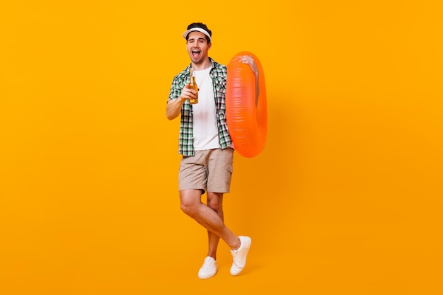 Free photo brunette guy in cap winks and poses with inflatable circle fulllength portrait of man in shorts with bottle of beer