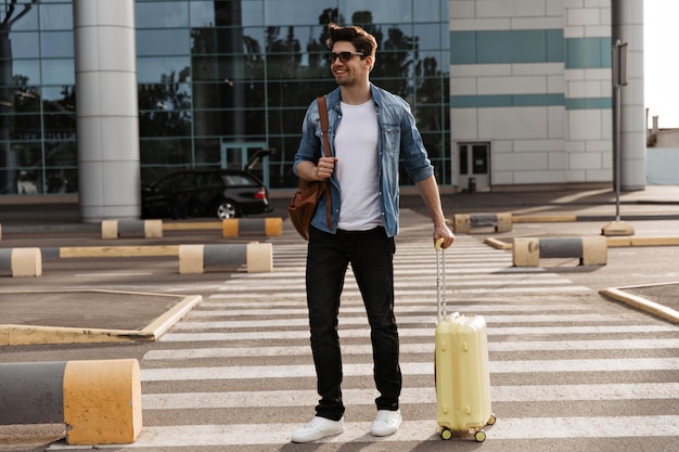 Free photo brunette handsome man in white tshirt and black pants walks on crosswalk cool guy in denim jacket and sunglasses holds brown backpack and yellow suitcase