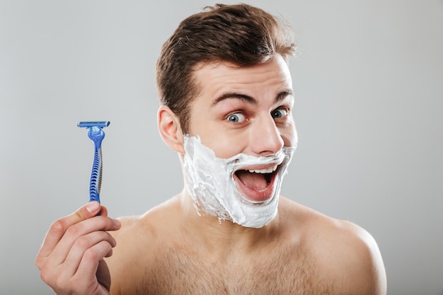 Brunette happy guy 30s being undressed at bathroom with shaving cream on face holding razor in hand over grey wall