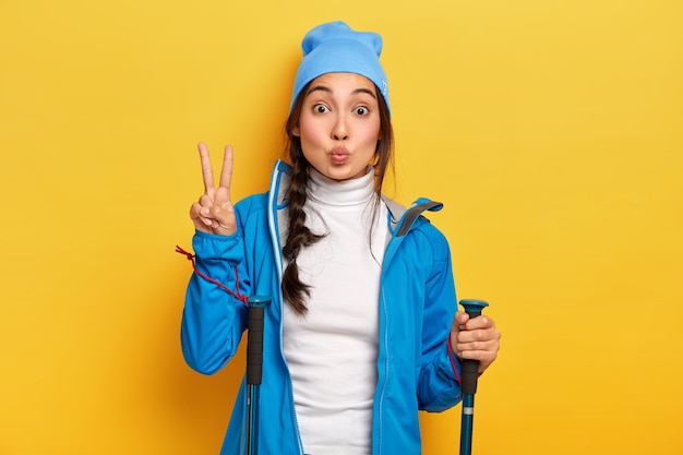 Brunette korean woman makes peace gesture, hikes in forest, holds trekking poles, dressed in blue casual clothes, enjoys active rest, poses over yellow wall