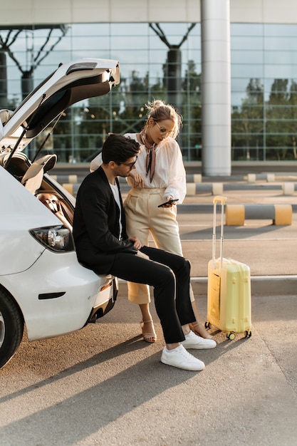 Brunette man in black suit and white t-shirt sits in car trunk