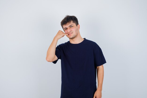 Brunette man holding finger in mouth in dark t-shirt and looking dissatisfied , front view.