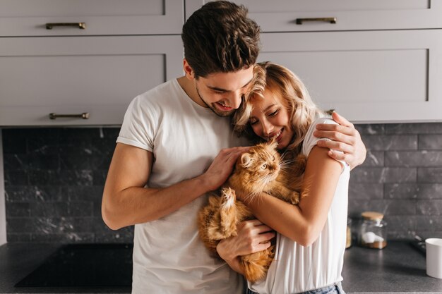 Brunette man looking at his cat and embracing wife. Indoor portrait of happy family posing with pet.