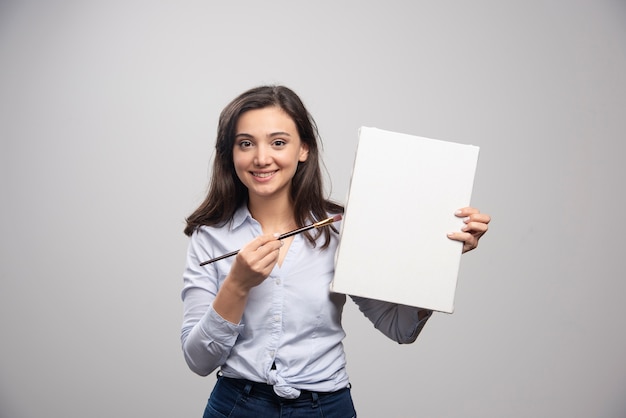 Brunette painter posing with canvas and brush on gray wall. 