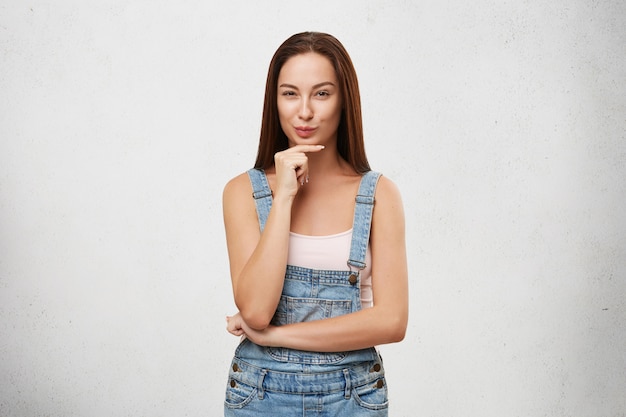 Brunette pretty woman wearing denim overalls, keeping hand on chin, looking with some intention to do something, having tricky plan in her mind.