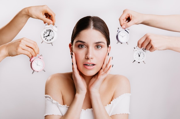 Brunette in white top closes her ears from alarm bell. Portrait of girl with green eyes on white wall.