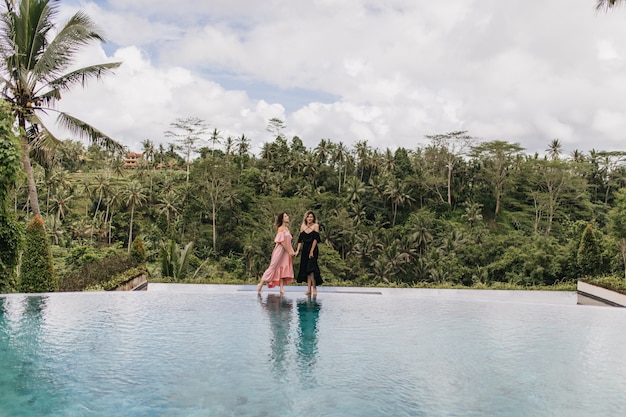 Free Photo brunette woman in pink dress holding hands with friend in bali. outdoor photo of female models standing near pool on jungle.