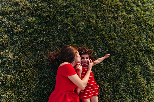 Brunette woman in red dress lying on the grass and kissing daughter in cheek. Outdoor overhead portrait of young mother and her kid chilling in park.