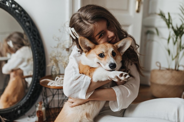 Free photo brunette woman smiling and hugging corgi happy young curly lady in white blouse has fun with cute dog in cozy room