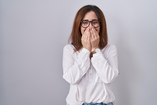 Brunette woman standing over white isolated background laughing and embarrassed giggle covering mouth with hands gossip and scandal concept