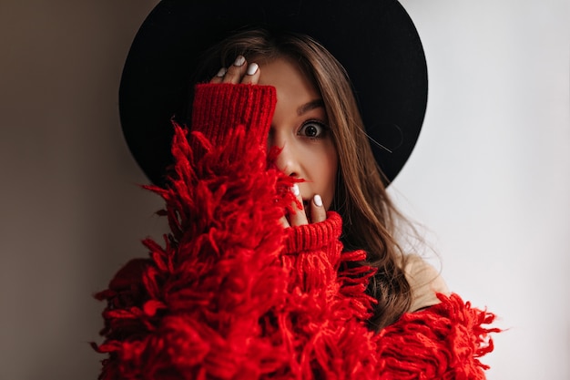 Free photo brunette woman in wide-brimmed hat looks at camera in amazement and covers her face with her hands. portrait of lady in knitted jacket.