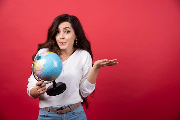 Brunette woman with globe feeling puzzled on red background. High quality photo