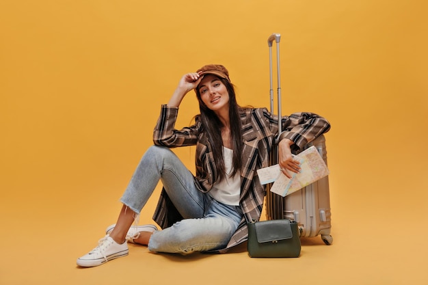 Free photo brunette young woman in beret white tee coat and jeans looks into camera and smiles stylish girl sits on floor leans on suitcase and holds map on isolated yellow background