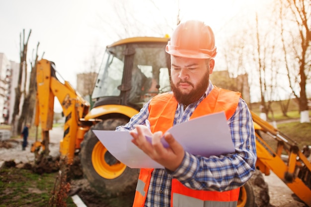 Free Photo brutal beard worker man suit construction worker in safety orange helmet against traktor with plan paper at hands