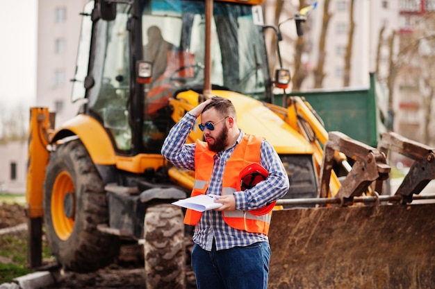 Free Photo brutal beard worker man suit construction worker in safety orange helmet sunglasses against traktor with plan paper at hands