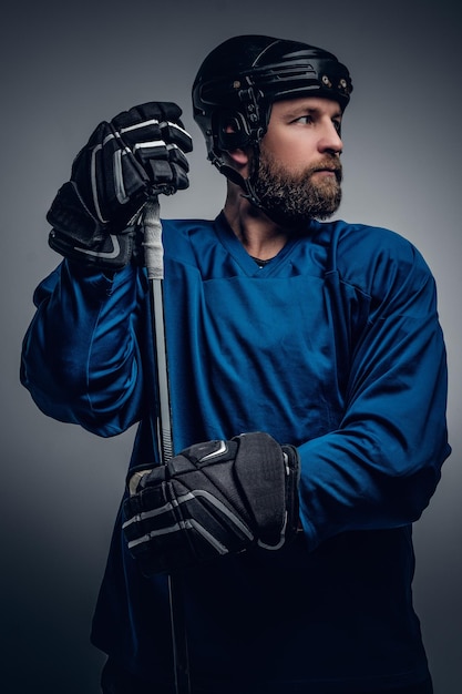 A brutal bearded ice-hockey player in safety helmet holds the gaming stick on grey vignette background.