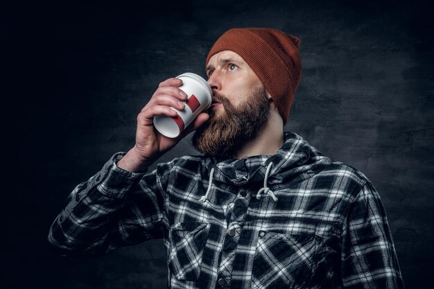 A brutal bearded male dressed in a hat and fleece shirt, drinks coffee from a paper glass.