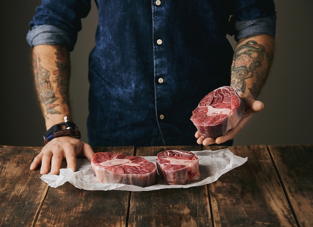 Free photo brutal butcher with tattooed hands offers a piece of great raw meat steak on camera, other steaks in white craft paper on vintage old wooden grunge table. unrecognizable