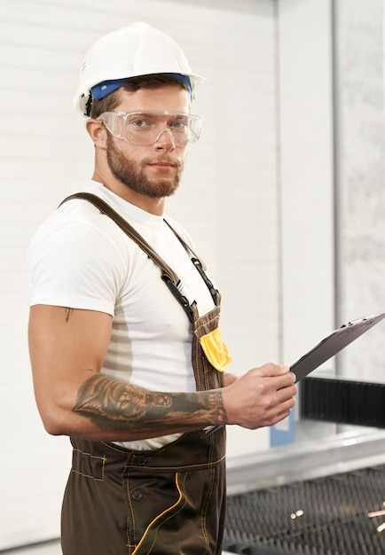 Free photo brutal engineer in uniform holding folder, posing on factory