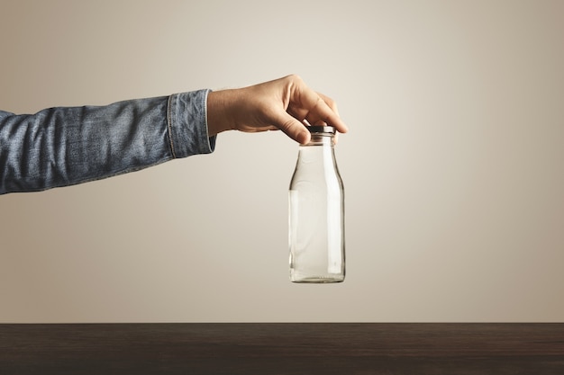 Brutal hand in jeans jacket holds glass transparent bottle with clean drinking water for black metallic cap above red wooden table, isolated on white