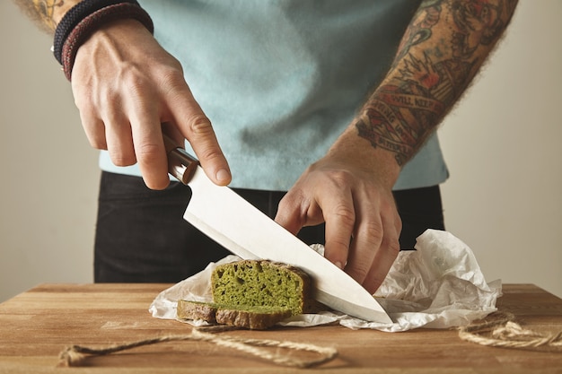 Brutal man tattooed hands cut healthy spinach home made green rustic bread with vintage knife on slices. Wooden board white table