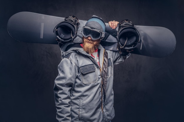 Free photo brutal redhead snowboarder with a full beard in a winter hat and protective glasses dressed in a snowboarding coat holds a snowboard on his shoulder in a studio. isolated on gray background.