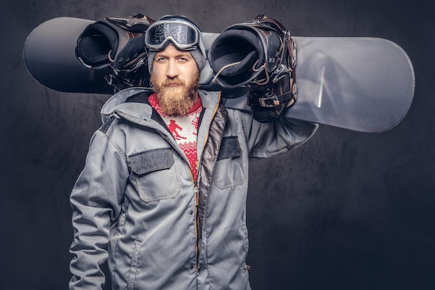 Brutal redhead snowboarder with a full beard in a winter hat and protective glasses dressed in a snowboarding coat holds a snowboard on his shoulder in a studio. Isolated on gray background.