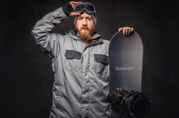 Free photo brutal redhead snowboarder with a full beard in a winter hat and protective glasses dressed in a snowboarding coat posing with snowboard at a studio. isolated on a gray background.