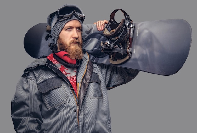 Brutal redhead snowboarder with a full beard in a winter hat and protective glasses dressed in a snowboarding coat posing with snowboard at a studio, looking away. Isolated on a gray background.