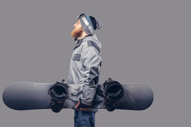 Free photo brutal redhead snowboarder with a full beard in a winter hat and protective glasses dressed in a snowboarding coat posing with snowboard at a studio, looking away. isolated on a gray background.