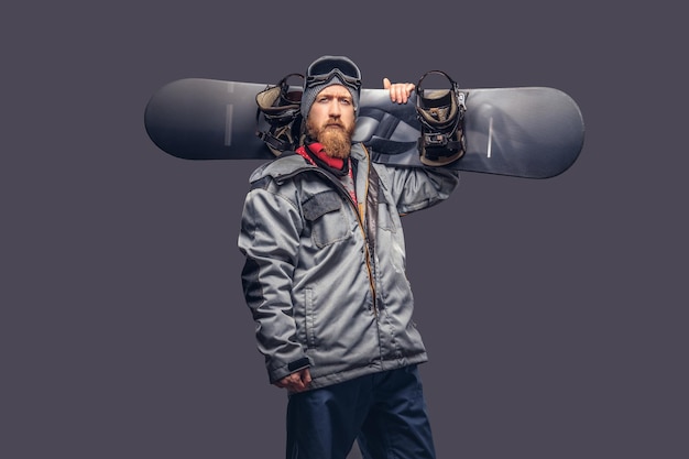 Free photo brutal redhead snowboarder with a full beard in a winter hat and protective glasses dressed in a snowboarding coat posing with a snowboard in a studio, looking at a camera. isolated on a gray backgrou