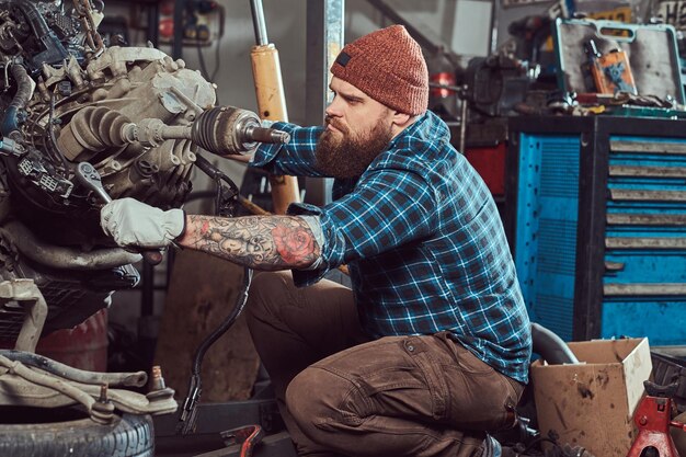 Brutal tattooed bearded mechanic specialist repairs the car engine which is raised on the hydraulic lift in the garage. Service station.