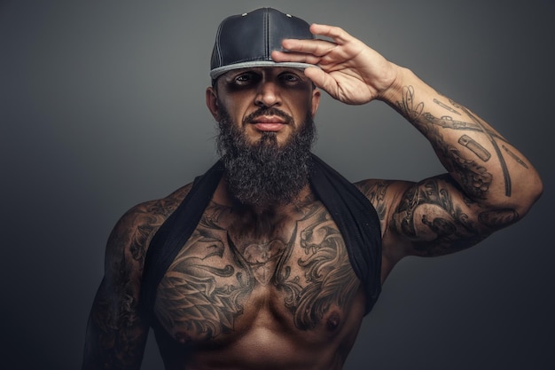 Brutal tattooed man in black cap posing in studio. Isolated on grey background.