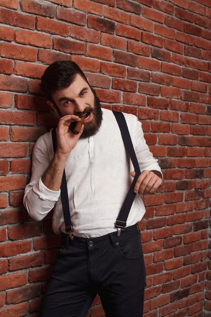 Brutal young handsome man smoking cigar on brick wall.