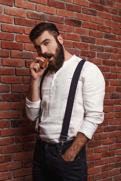 Brutal young handsome man smoking cigar on brick wall.